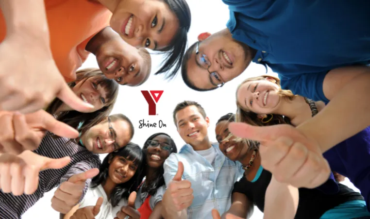 A group of young people are looking down toward the camera and are smiling and holding their thumbs up. The YMCA Shine On logo is in the middle