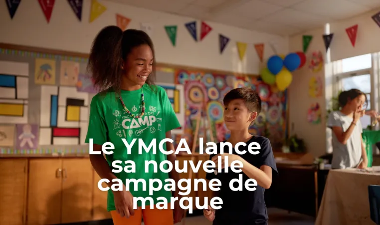 Texte : LE YMCA LANCE SA NOUVELLE CAMPAGNE DE MARQUE. Une fille et un garçon se tiennent debout dans une salle de garderie en souriant.